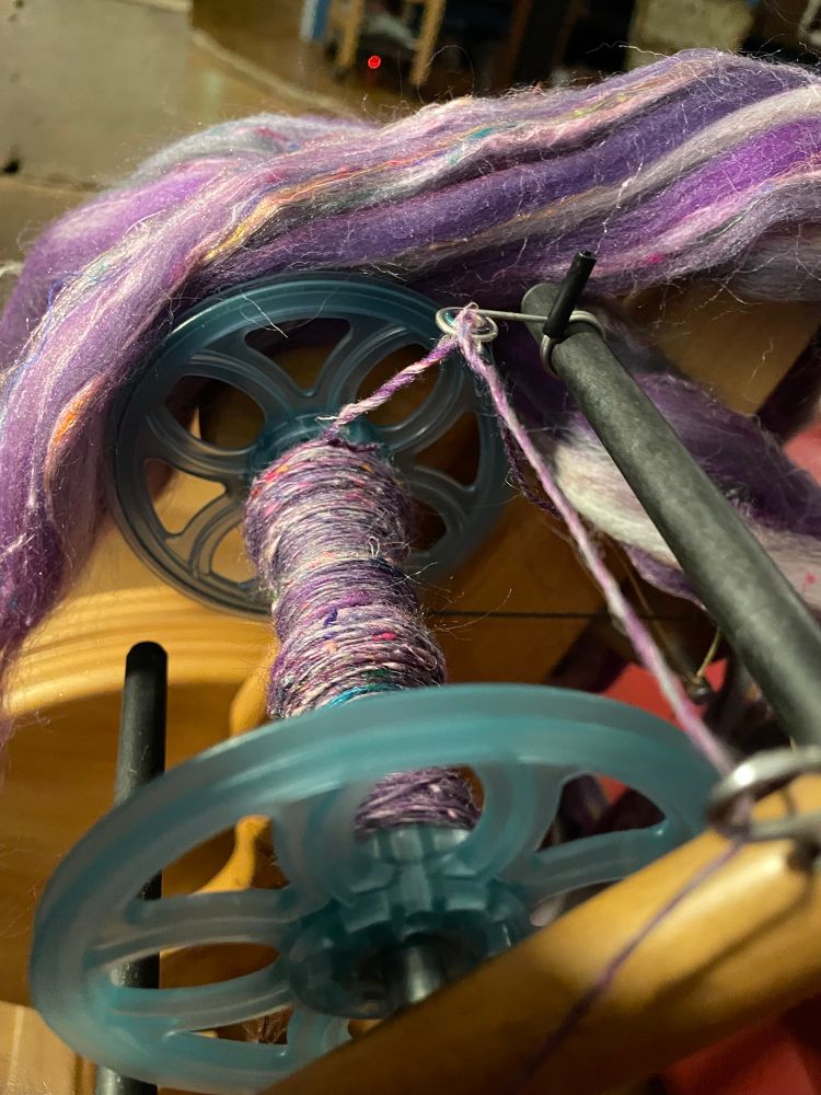 Spinning in progress, closeup of a bobbin with purple wool mixed with little colourful sat silk bits, the combed top is draped over the wheel. 