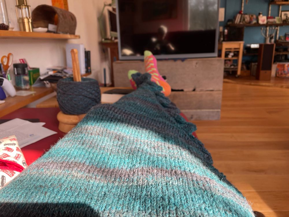A knitting project in teal and grey/black colours spread over outstretched legs, covering them. A big ball of the handspun yarn is in the background to the left. 