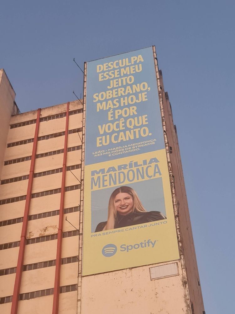 Outdoor gigante do spotify na lateral de um prédio com o trecho da música "Desculpa esse meu jeito soberano, mas hoje é por você que eu canto" da cantora Marilia Mendonça