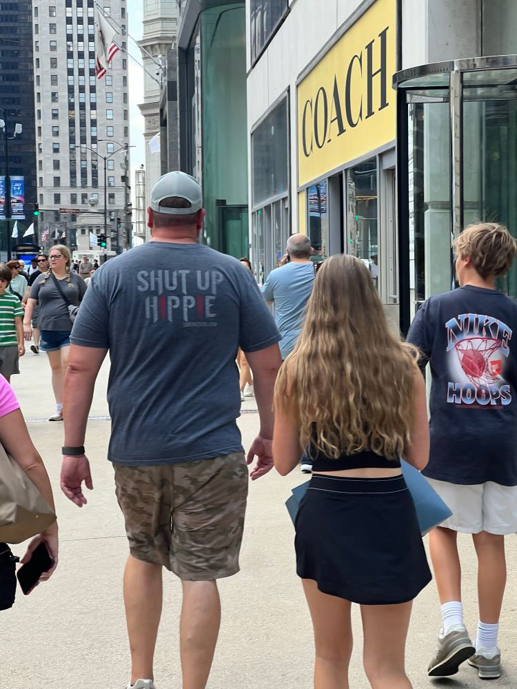 A dad in camo shorts and a t-shirt escorts his family past the hair-raising danger of the entrance of a Coach store. The back of his t-shirt reads “shut up hippie” — with each ‘I’ in the last word marked with red-coated, downward pointing bullets. 

The rest of his family seems to be enjoying a normal day, like normal people. 