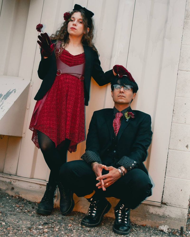 A shot of a woman in a red dress and black blazer holding a red flower in front of a white wall. Crouched beside her is a man in a black suit and red tie, and her red gloved hand rests atop his head.