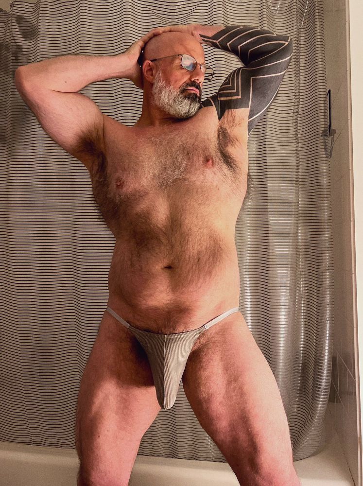 A hairy dad in a bathroom with his hands clasped behind his head, exposing his musky, hairy armpits. He’s wearing a pair of striped briefs that’s being stretched by the meaty object inside. 
