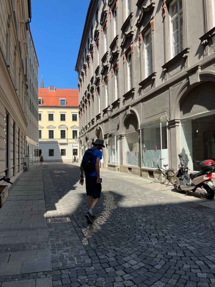 A man with a hat and a rucksack walking along a typical looking European town centre cobbled street. In the middle is a line of some shiny bronze cobblestones resembling the effect of a pathway thats been worn away by multiple feet on the same shortcut