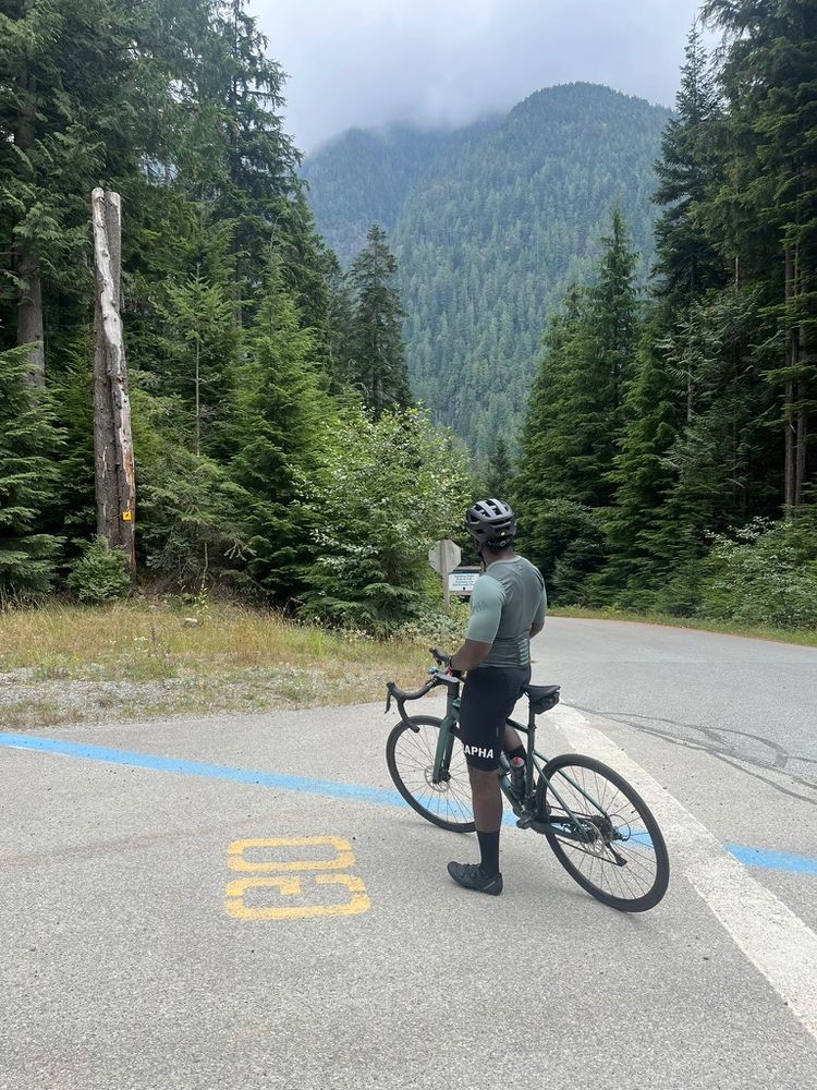 cyclist with bike standing on mountain road with forested hills in background.