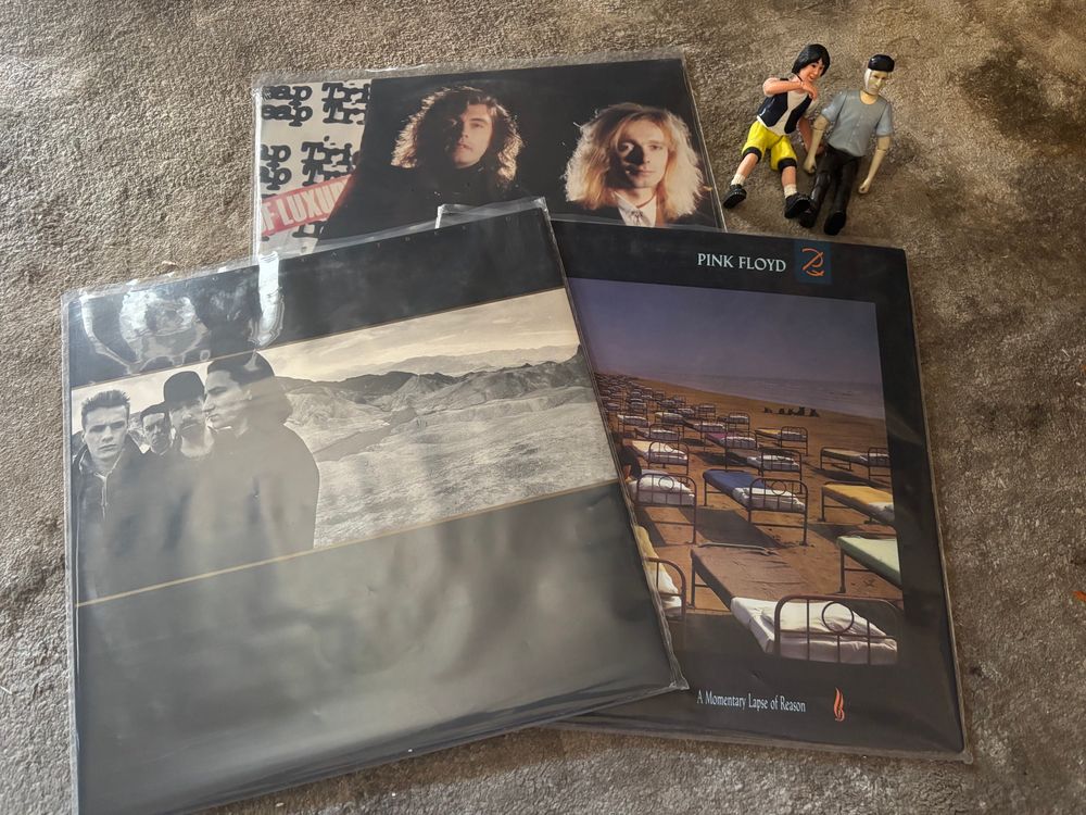Three vinyl albums scattered on top of each other in a triangle on the floor. Starting from the top and going clockwise they are:
Cheap Trick - Lap of Luxury 
Pink Floyd - A Momentary Lapse of Reason 
U2 - The Joshua Tree
Ted Theodore Logan and Vincent Van Gogh action figures are seated next toy the albums. 
