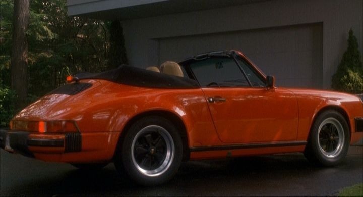 A red 1984 Porsche 911 convertible with tan interior parked in the driveway outside of a home garage.  The photo angle is the passenger side slightly angled from the back. 
A scene from the movie Opportunity Knocks.