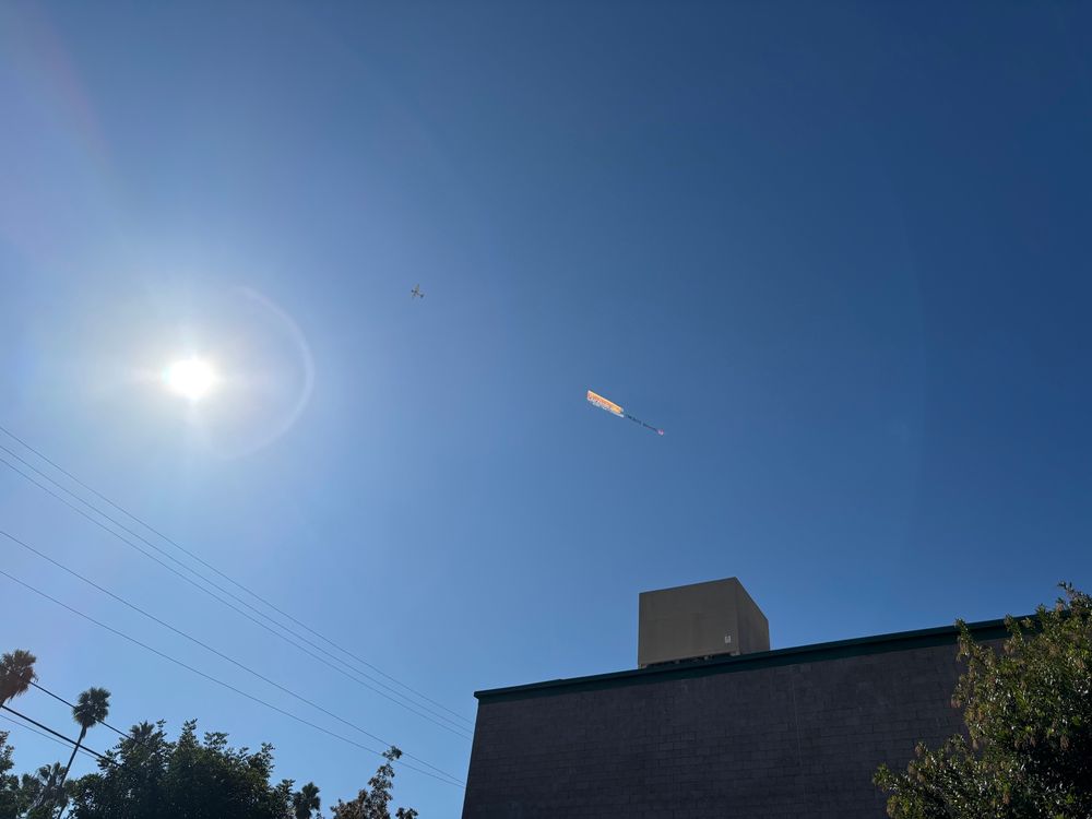 A small airplane in the clear blue sky towing a banner behind it. To the left is the sun and below is a building and some tree tops. 