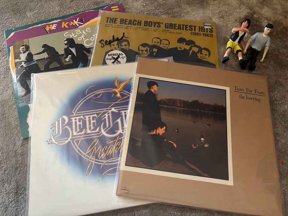 Four vintage vinyl records scattered on top of each other on the floor. Starting top left and going clockwise they are:
The Kinks - State of Confusion 
The Beach Boys’ Greatest Hits 1961 to 1963
Tears For Fears- The Hurting
Bee Gees Greatest 
Ted Theodore Logan and Bincent Van Gogh action figures are seated next to the albums. 