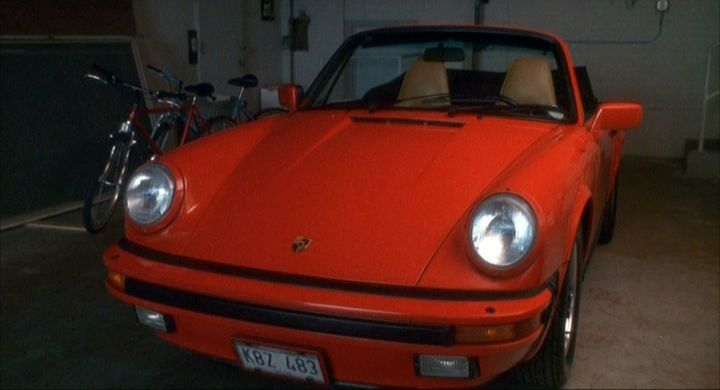 A red 1984 Porsche 911 convertible with tan interior parked in a home garage.  The photo angle is the front driver’s side corner. 
A scene from the movie Opportunity Knocks.