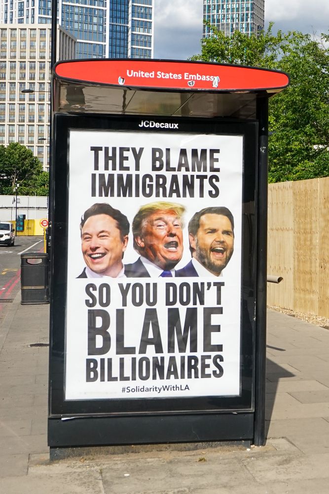 A guerilla bus stop advert outside the US embassy in London. It features pictures of Elon Musk, Donald Trump and JD Vance. The text on the ad reads 'they blame immigrants so you don't blame billionaires'