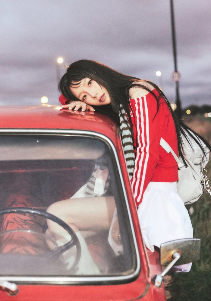 Taeyeon perched on the driver side window of a red car, her head resting on the top of the car. She wears an off shoulder red top, white skirt, and a backpack.