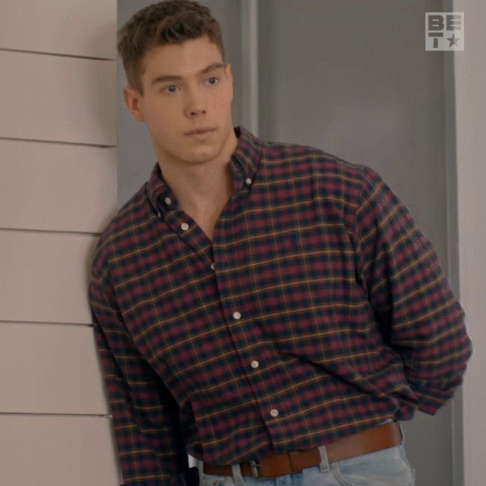 a man in a plaid shirt leans against a wall with a be tv logo in the corner