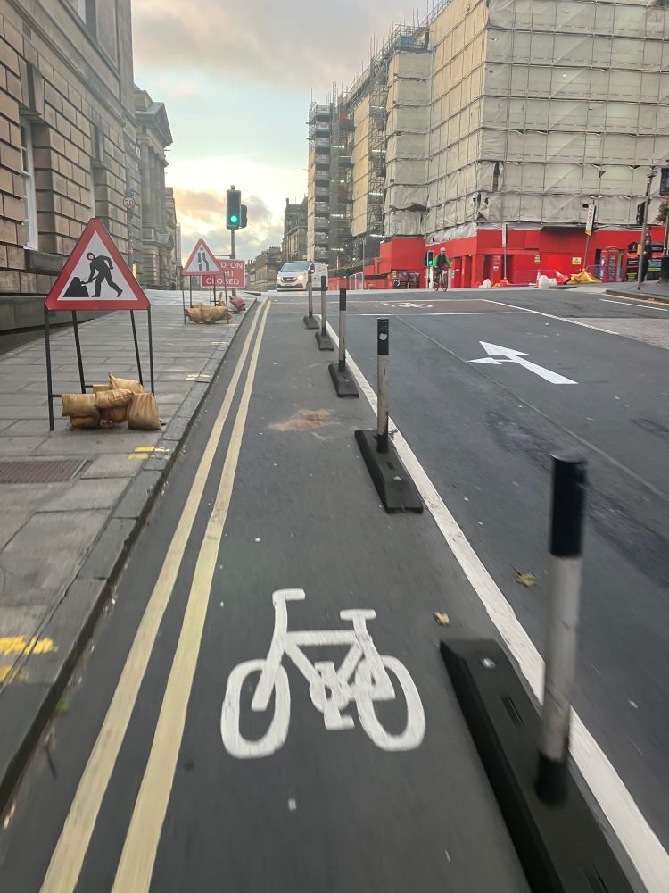 A protected cycle lane with temporary road signs on the pavement to the left and a clear wide road to the right. There is a junction ahead with traffic lights at green