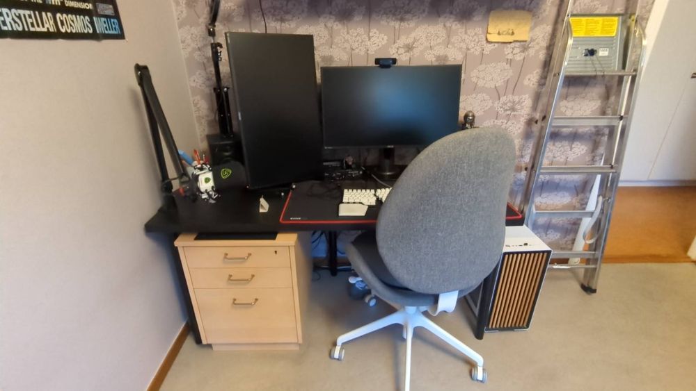 Foto av ett trångt datorbord som med nöd och näppe rymmer två stora skärmar, två högtalare och en hållare för en mikrofon. Bordet är svart. Under bordet står en hurts och en datorstol står framför det och ser ut att vara gigantisk jämfört med bordet.