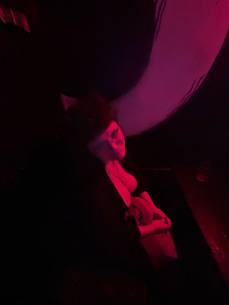 A dark photo of an alt femme nonbinary person sitting at a bar table in red lighting, and they look good as fuck