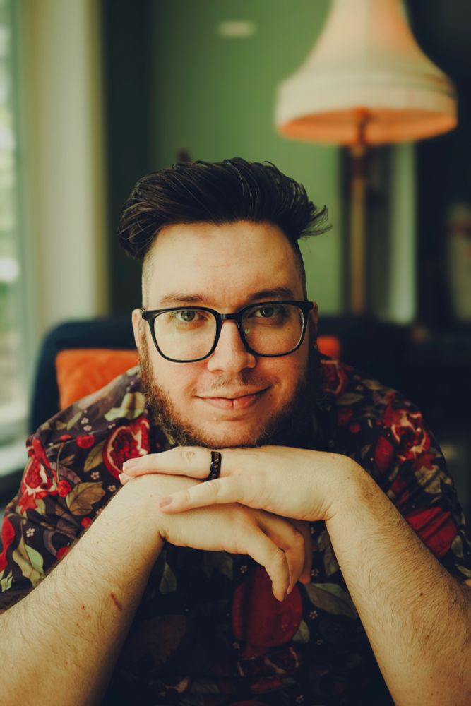 my gorgeous husband posing for a photo. he’s wearing a colorful goth tee and sitting with his hands under his chin. 