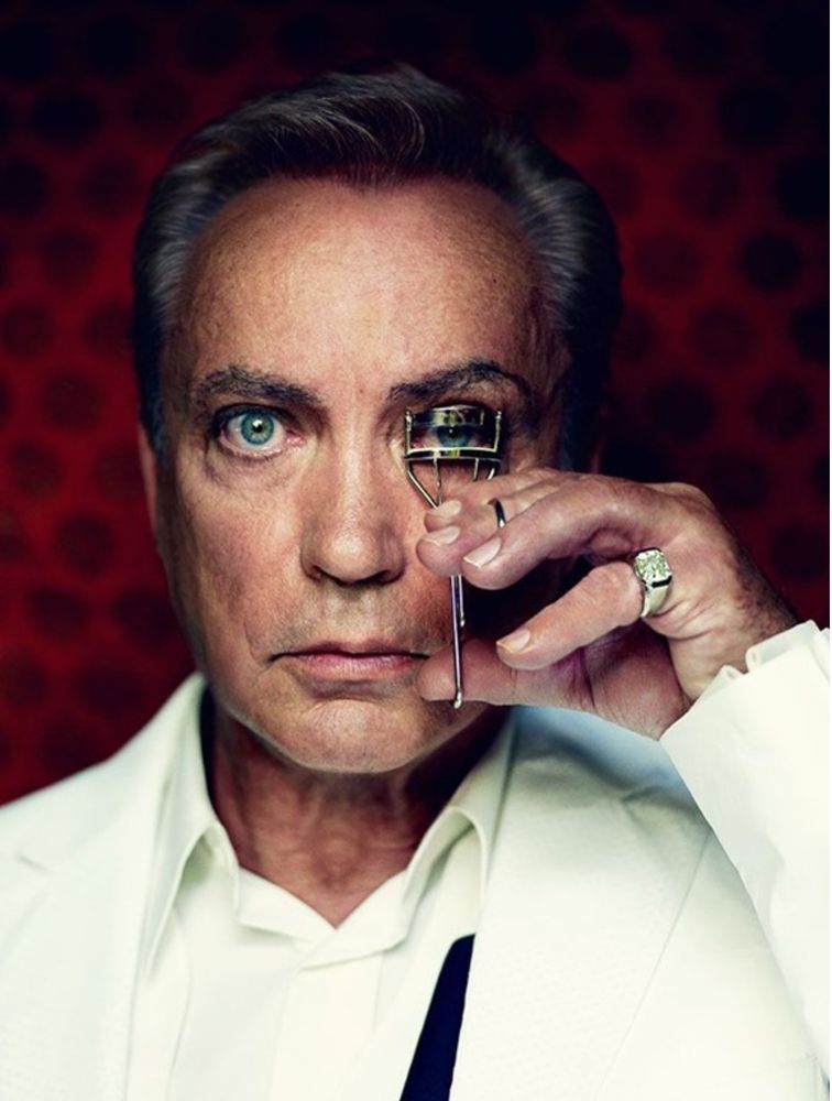 German actor Udo Kier in a super crisp white shirt and jacket holds an eyelash curler to his left eye while his bright blue right eye Pierce’s your soul
