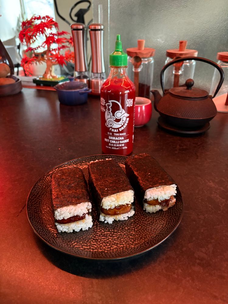 Three spam musabi on a black plate on a black kitchen counter, a bottle of sriracha behind it