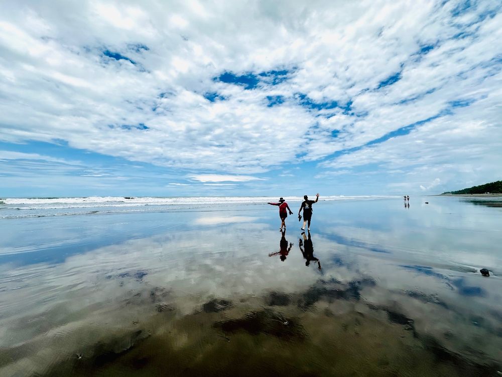 Photograph of a beach covered with a thin layer of water. The water is reflecting the white clouds and blue sky.  Two people are walking and waving.