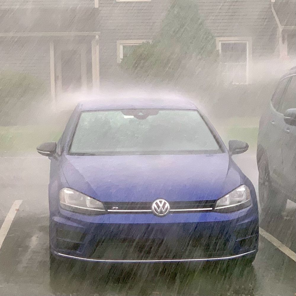 The front of blue VW Golf parked in a parking spot in a heavy rainstorm, with rain being blown diagonally from right to left 