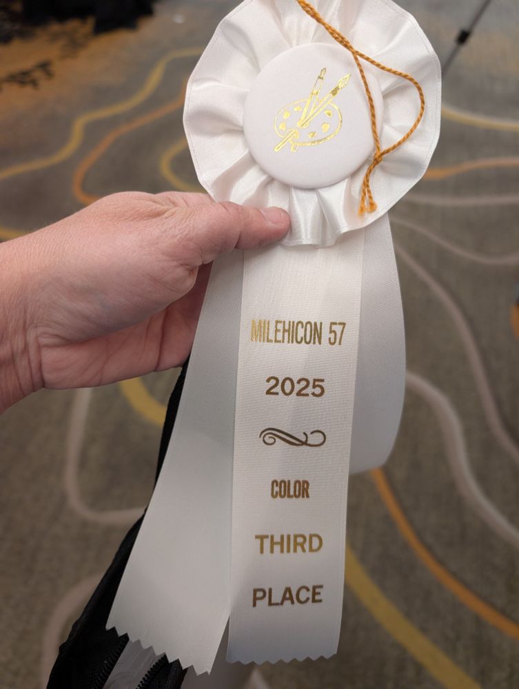 A hand holding a white winner's ribbon that says "Mile Hi Con 57, 2025, Color Third Place"