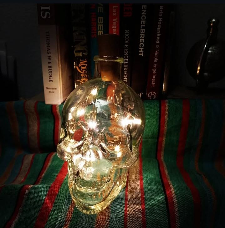 Glass skull shaped bottle containing fairy lights on a striped cloth and a selection of fiction and nonfiction books in the background.
