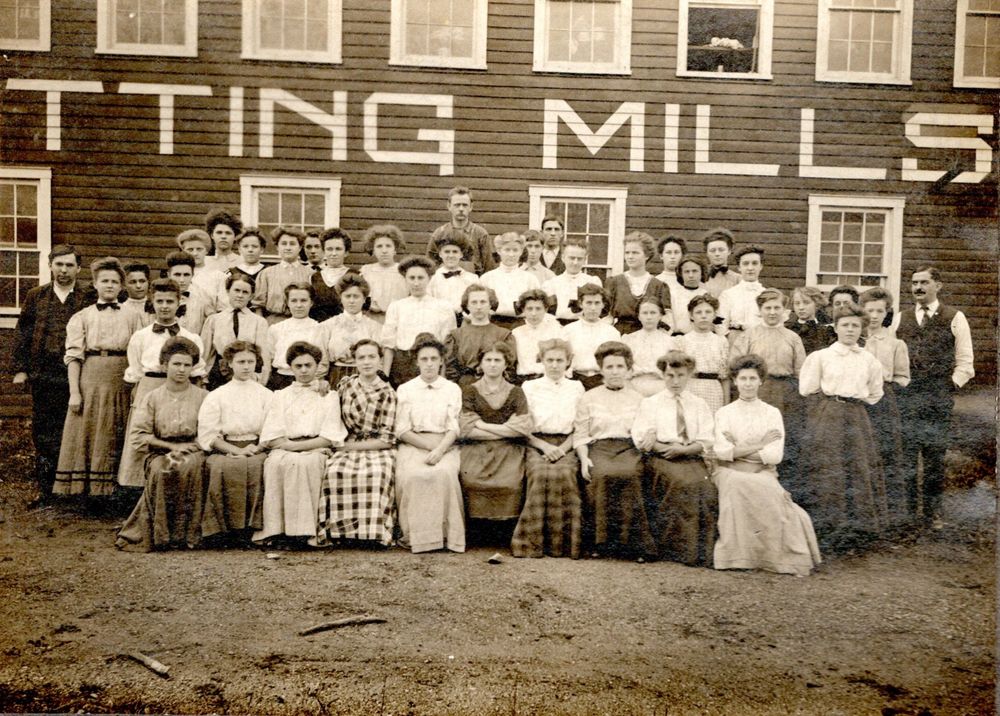 A group of about 50 women and 5 men arranged outdoors in 4 rows. The first row of 10 women is seated. Everyone else is standing, all in front of a dark colored clapboard mill building fitted with closely set 6 over 6 windows. One window is open. Large text is painted on the building. The text that’s visible reads “TTING MILLS” The women are all quite young, some just barely in their teens. Many of the women wear light colored blouses with elbow length sleeves and long dark skirts. Several of the women are wearing large coverall aprons. The 3 men at the center back appear to be wearing work clothes. A young man on the left wears a waistcoat and jacket but no collar or tie. An older man on the right wears a waistcoat over a light shirt worn with a tie and a stiff collar. He is looking to his right (possibly trying to spot the person who just giggled)