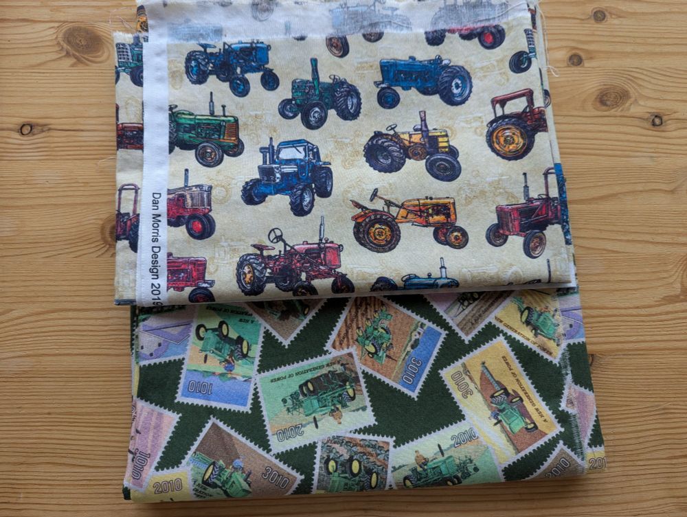 Two folded up pieces of fabric. One has vintage tractors in blues, greens, oranges, and reds against a cream background. The other has small rectangular pictures of vintage John Deere tractors against a green background 
