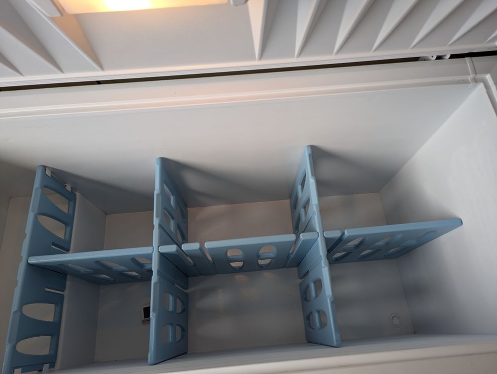 The white interior of a 19 cubic foot chest freezer. Pale blue dividers have been installed, dividing the space into sixths so that there is the ghost of a chance that the chicken can be segregated from the pound cake and the pound cake from the sausage. This sort of works.