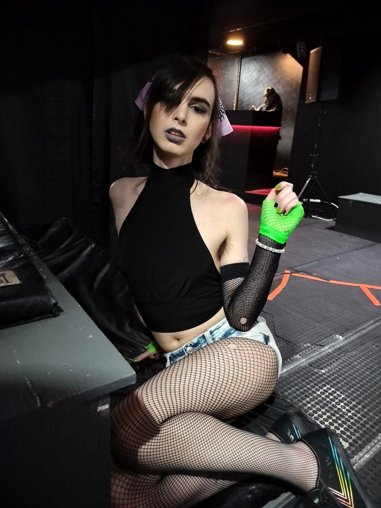 white trans woman in a black sleeveless and cropped shirt, fishnets with jean booty shorts, fishnet arm sleeves and a purple bandana.