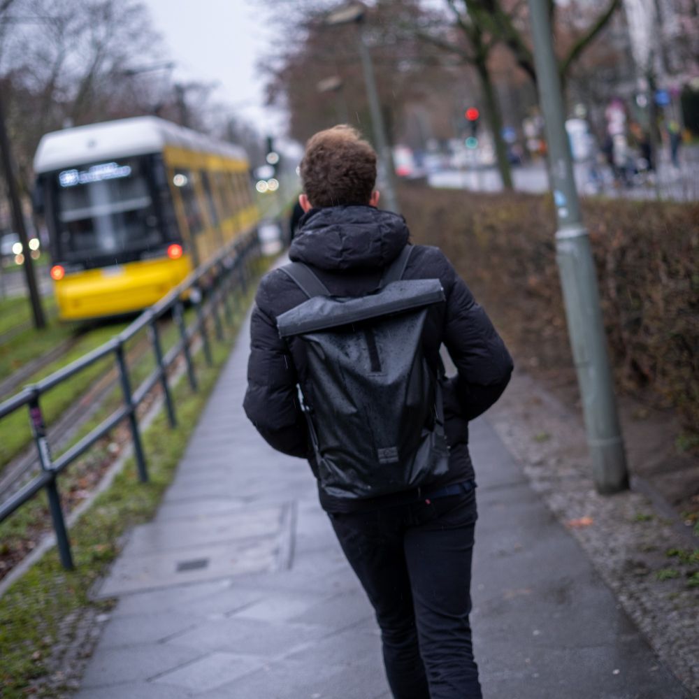 Gehend auf dem Weg an der Straßenbahnhaltestelle am U-Bahnhof Seestraße in Richtung Cornelius-Fredericks-Straße. Im Hintergrund ist die abfahrende Straßenbahn der Linie M10 zu erkennen. Kurz zuvor hat es geregnet, es sind noch feuchte Stellen auf dem Boden zu erkennen.