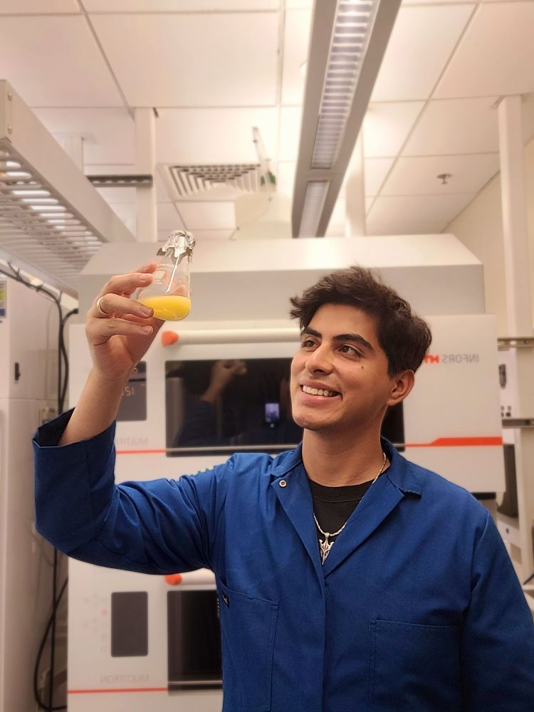 A researcher in a blue lab coat holds up a beaker with yellow liquid in it. 