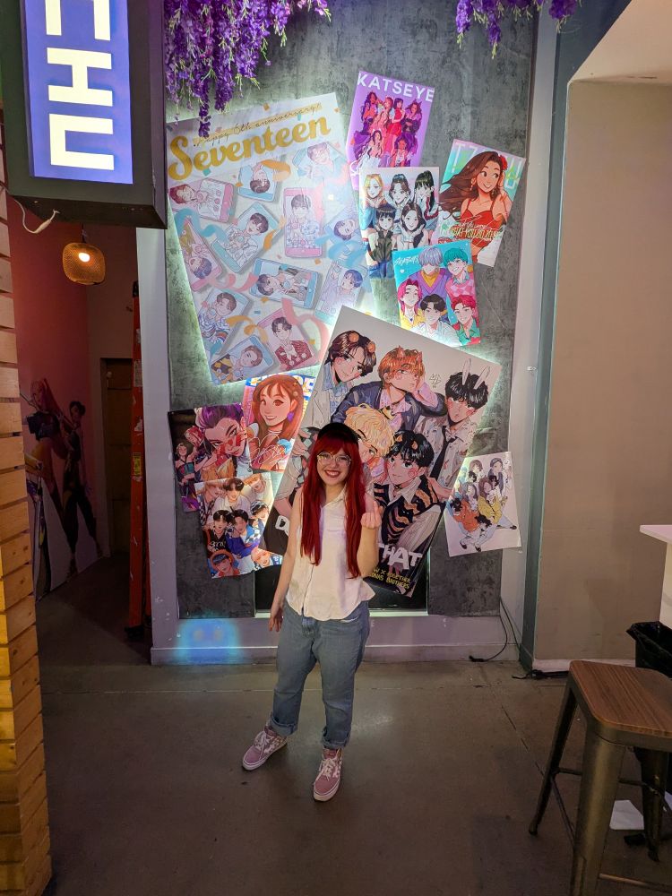 Me cosplaying as Mira from Kpop Demon Hunters in her casual/undercover outfit when they first see the Saja Boys performing Soda Pop. I'm at the Pop Fancy KPDH pop up cafe, posing in front of their wall of Kpop posters and making a heart with my fingers