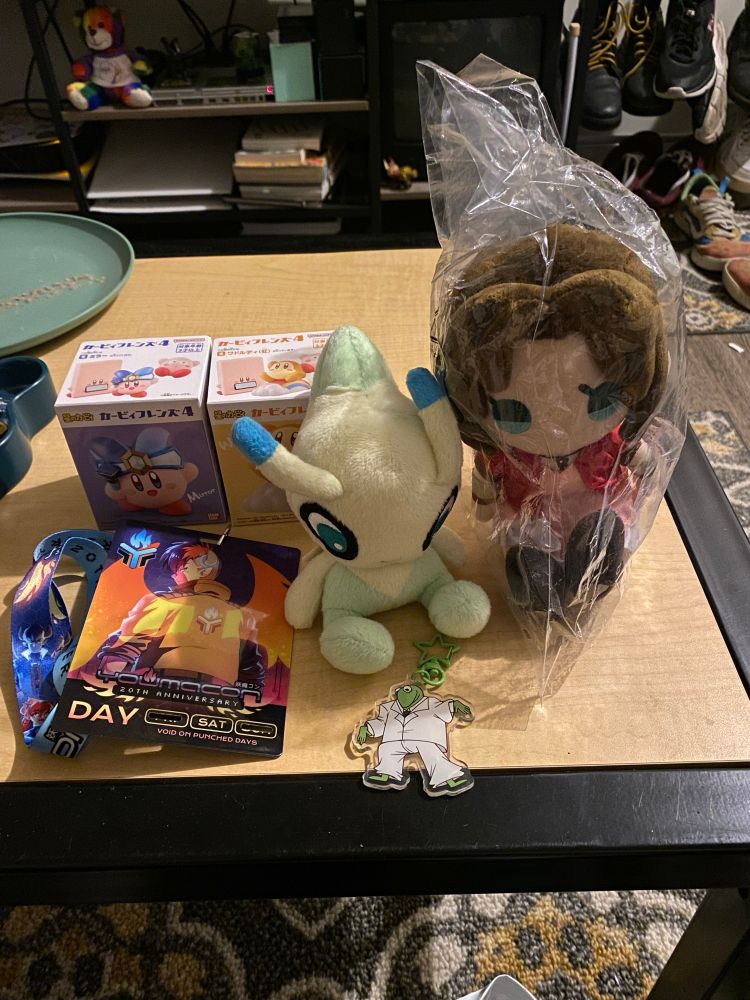 A collection of items from Youmacon 2025, including the badge, two Kirby figures, a Celebi plush, an Aerith plush, and a Kermit charm