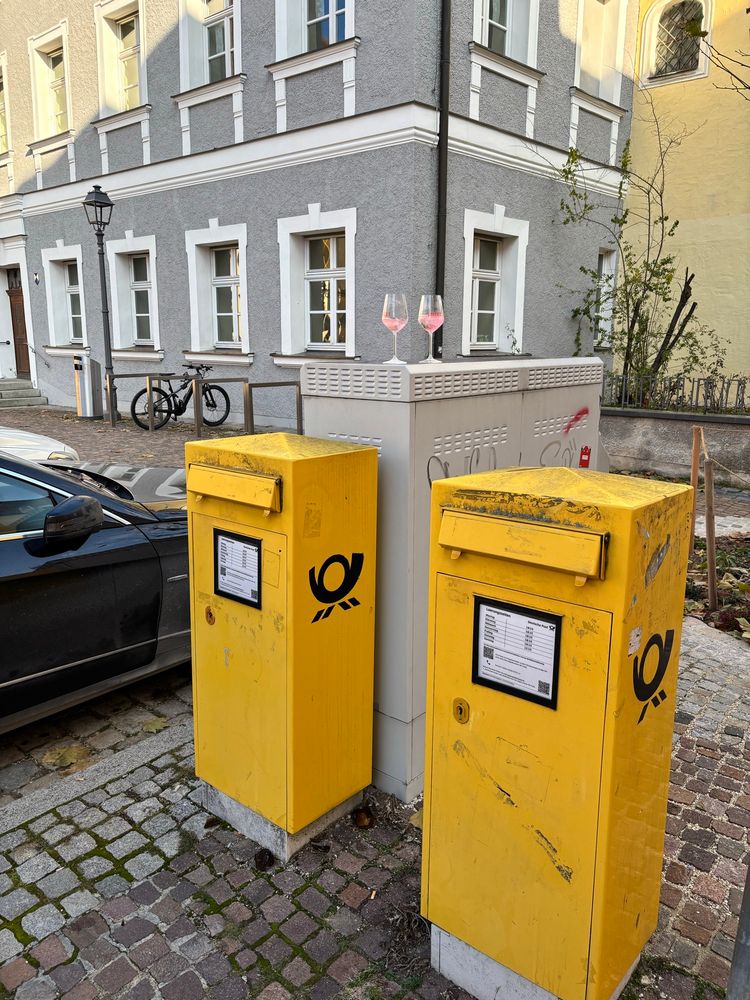 Zwei Lillet-Gläser, halb gefüllt, auf einem Kasten hinter zwei gelben Briefkästen am Straßenrand 