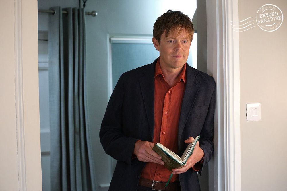 DI Humphrey Goodman leaning in a doorway, holding a book, and smiling.