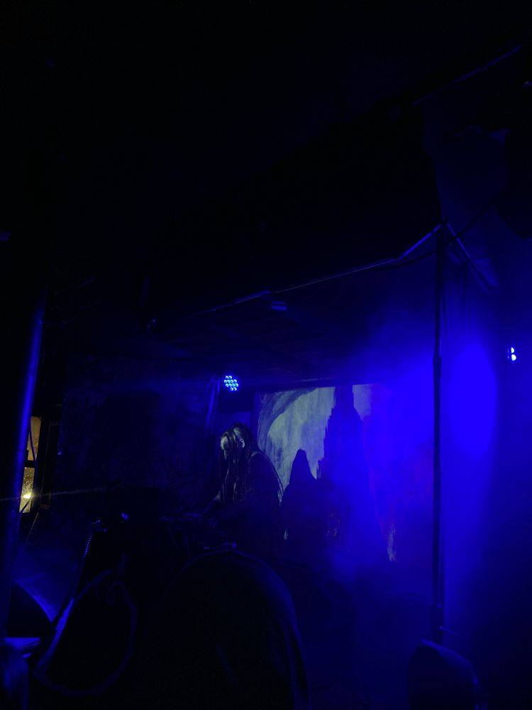 A performer on stage in fog, dressed in black and wearing a goblin mask, over a synthesiser. Behind them a projected image of a monochrome pencil drawing of a great black obelisk 