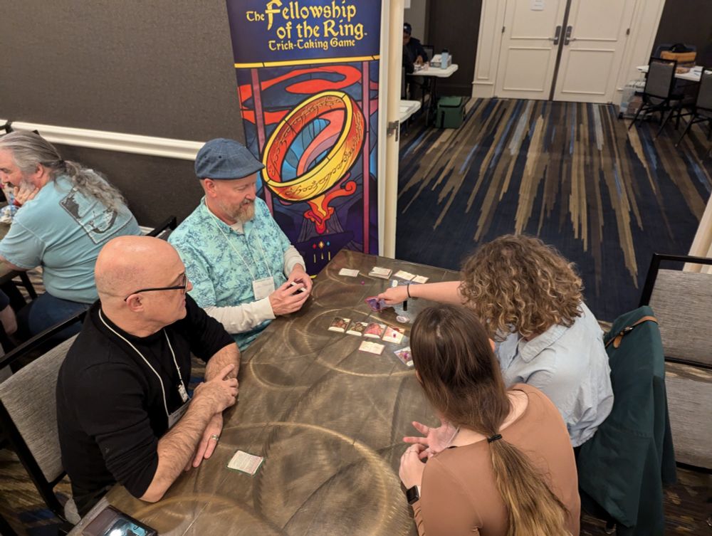 Luke Peterschmidt seated in front of a banner reading The Fellowship of the Ring Trick Taking Game at a table with three other players, learning the card game. 