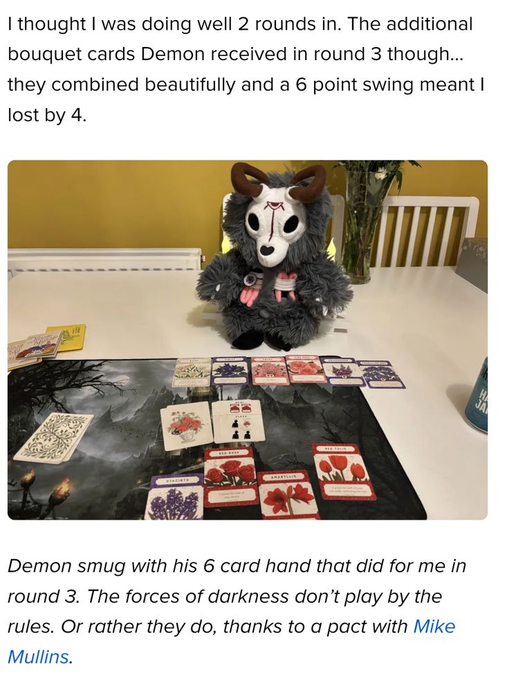 Screenshot of a BGG blog post. The text above reads, "I thought I was doing well 2 rounds in. The additional bouquet cards Demon received in round 3 though... they combined beautifully and a 6 point swing meant I lost by 4."

The image shows a stuffed demon serving as the opponent in a solo game of Tussie Mussie, with beautifully illustrated flower cards distributed on the table. 

The text below reads, "Demon smug with his 6 card hand that did for me in round 3. The forces of darkness don't play by the rules. Or rather they do, thanks to a pact with Mike Mullins."