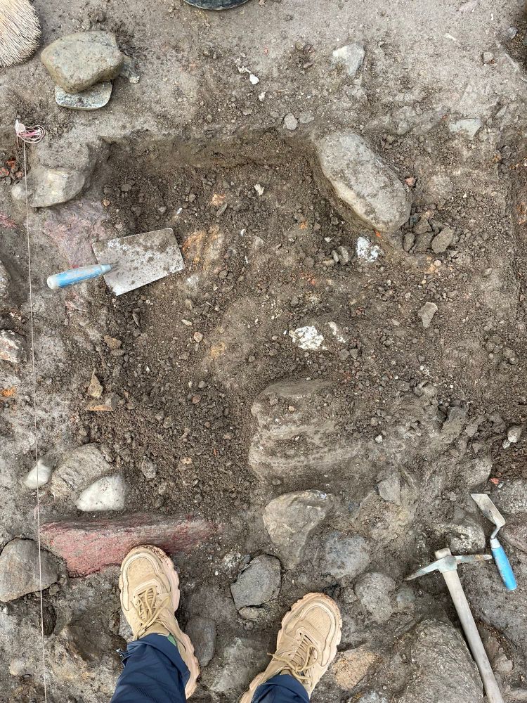 Schnitt auf der heurigen Grabungskampagne von oben. Zwei Füße in beigen Sicherheitsschuhen, Steine, Erde und Werkzeuge. 