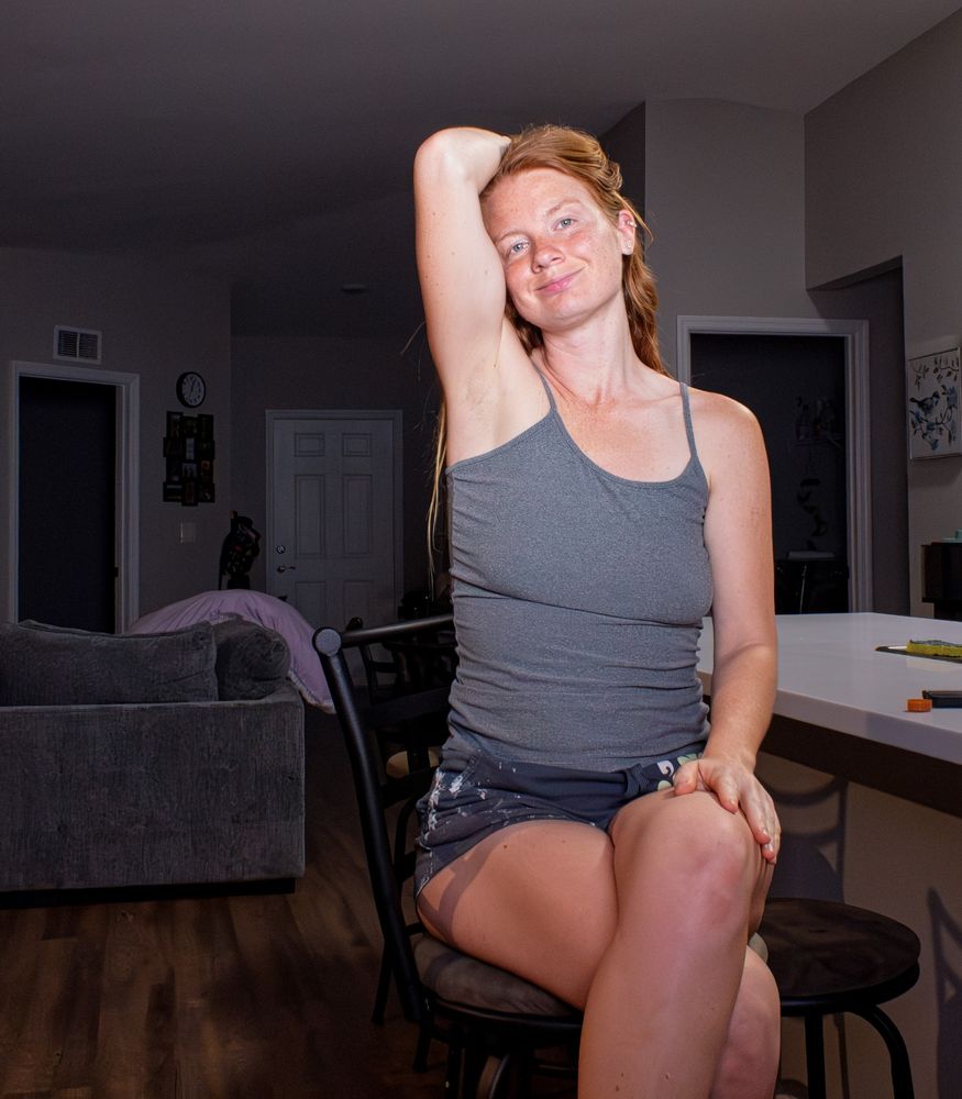 Lily Rose, a beautiful redheaded woman with long ginger hair and a face and arms full of freckles, sits on a kitchen bar stool. She smiles sweetly at the camera, resting one arm behind her head. She is wearing a grey tank top and dark shorts in the softly lit home interior.