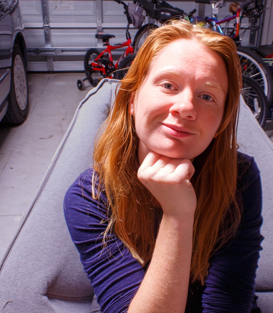 Lily Rose, a beautiful redheaded woman with long ginger hair and a face and chest full of freckles, is sitting on a gray inflatable couch or cushion in a garage. She is wearing a long-sleeved dark blue top and smiling softly at the camera, with a car and various bicycles visible in the background.