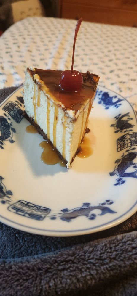 A slice of rich, decadent plain cheesecake on a white plate that's dressed with a cherry on top and a caramel sauce that drips down its sides