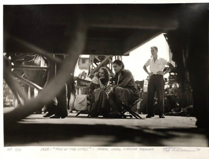 Ronald Colman and Isabel Jewel sit quietly and thoughtfully in costume, in folding chairs turned somewhat toward each other, outdoors on the set of A Tale of Two Cities. The photo has been taken from some distance away and your view passes underneath a shadowed wagon with large wooden wheels. You can see people milling about, at work, as Jewell and Colman stay to themselves. It's obvious they are shooting their last scenes of the film, which are emotional and dramatic for their characters.
BUT this version of the photo is taken from much further away, showing the much larger busier set around them and making them look much smaller in comparison to everything around them.