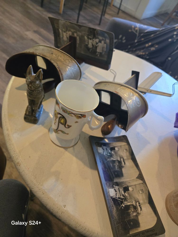 On a white coffee shop table are two stereoscopes (different manufacturers, but neat symbols), viewing pictures for stereoscopes, Bastet statue and Irish coffee cup (according to various lookups) 