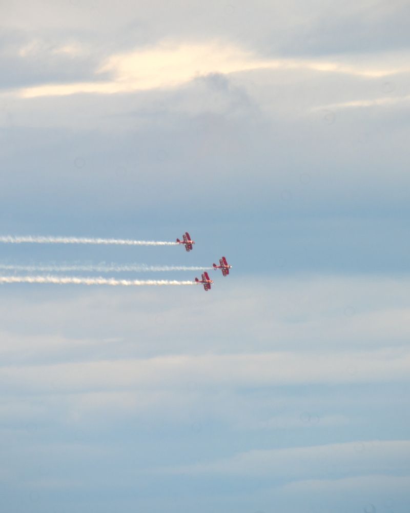 a trio of red and white biplanes fly in formation against a cloudy sky while trailing smoke