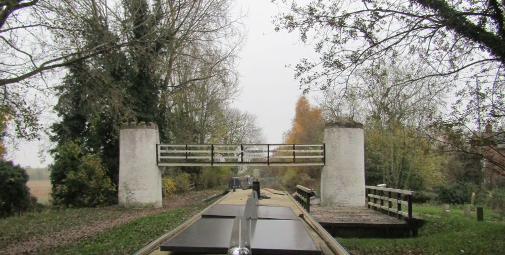 A photograph of an autumnal canal scene. The water is barely visible around the edges of a cream coloured boat taking up the lower centre of the frame. Over the boat is a white and black bridge supported on each end by white round turrets rimmed with stone castellations.
The towpath on the left is disappearing under fallen leaves, and on the right is a much lower, flatter, bridge pushed to one side of the canal. What sky that is visible is flat grey.