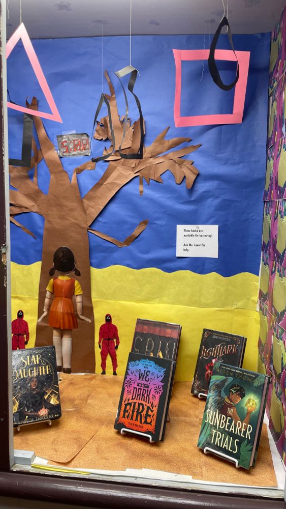 Images from the tv show Squid Game in the background of the display case: a doll viewed from reverse against a tree and 2 squid game guards. Books visible in foreground: Star Daughter, We set the dark on fire, Spin, Lightlark, and the Sunbearer Trials 