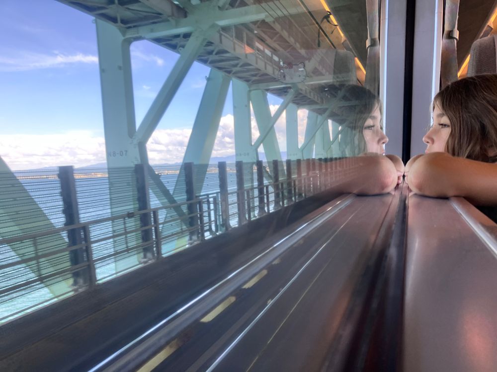 Young girl looking out window at bridge and ocean while her reflection looks back.