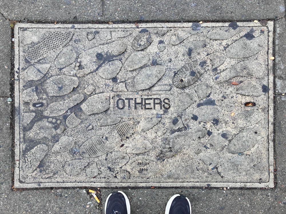 Sidewalk plaque with varied shoe soles and the word 'others' in the middle.  Toes of phototakers shoes stick into the picture